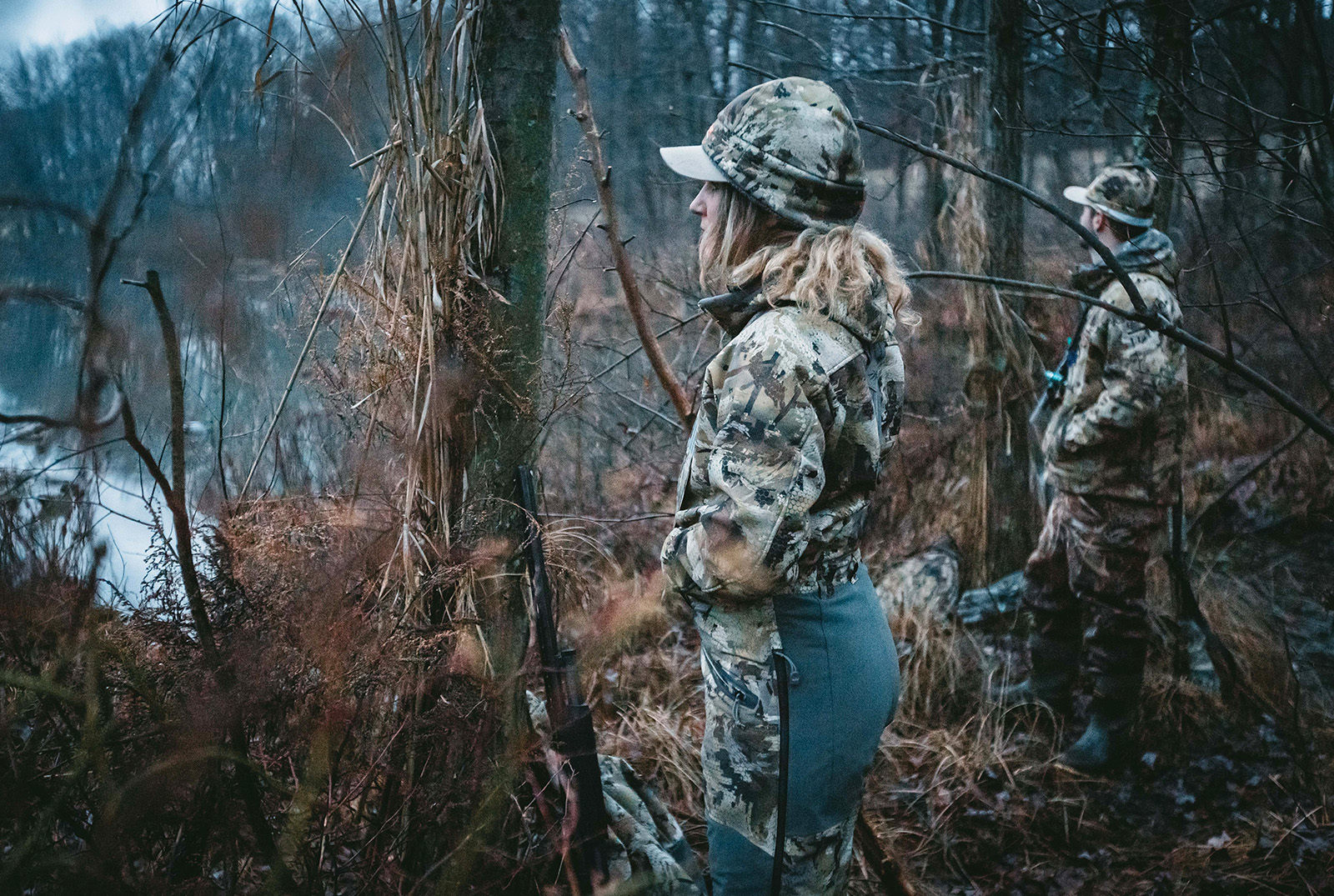 Two hunters look out onto a waterway.